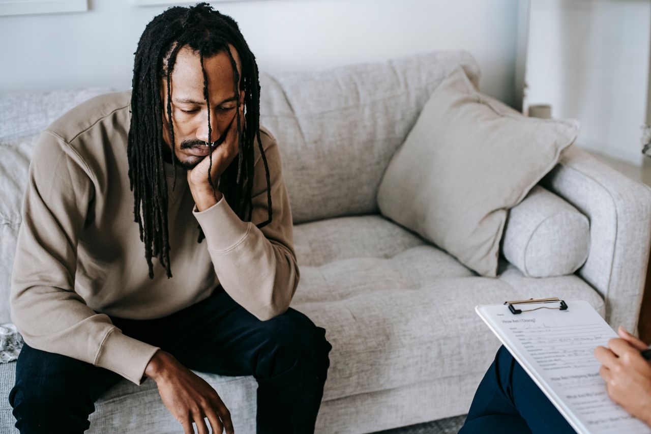 Upset African American male with dreadlocks leaning on hand and looking down while sitting on couch near crop psychologist with clipboard